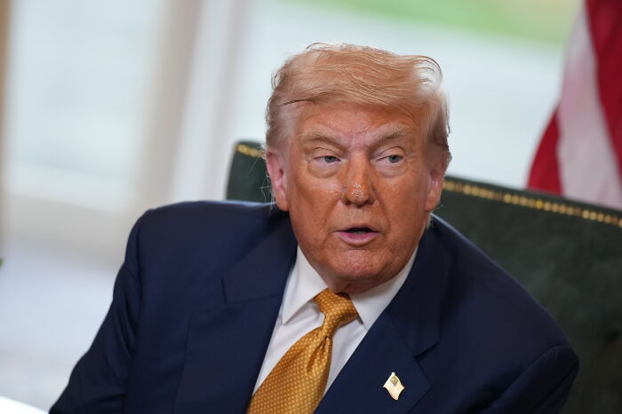 Former President Trump seated indoors wearing a navy suit and yellow tie discussing immigration and asylum seekers.