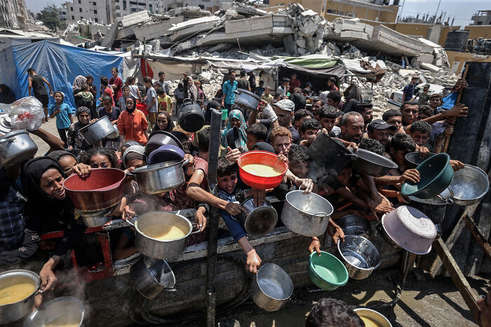 Crowd of people holding bowls and pots amid rubble in Gaza, highlighting the severity of the Gaza genocide situation.