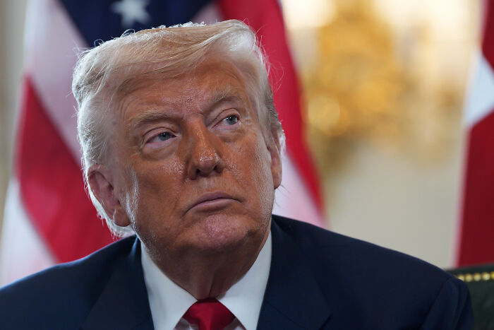 Donald Trump at a press briefing, looking serious with American flags and blurred background behind him.