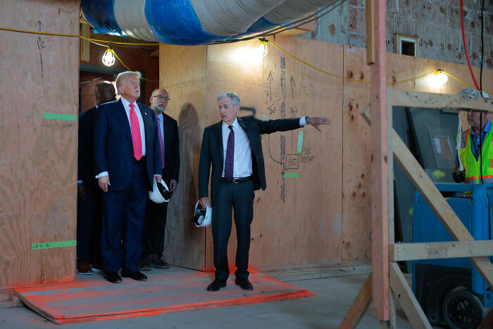 Federal Reserve Chair Jerome Powell in suit gesturing during a construction site visit with Donald Trump and others.