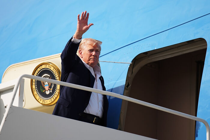 Donald Trump waving on Air Force One stairs with presidential seal in the background, related to Epstein and Mar-A-Lago.