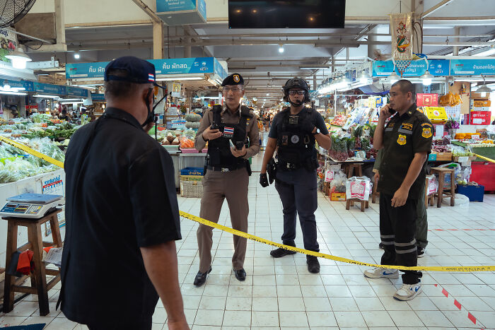 Police and security personnel at a food market in Bangkok investigating a mass shooting incident with yellow caution tape barriers.