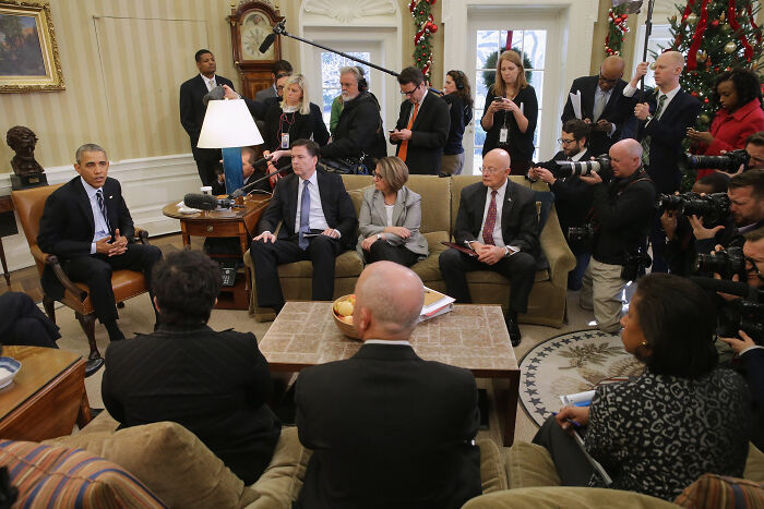 Barack Obama seated in a meeting room with media and officials capturing the session on cameras and recording devices.