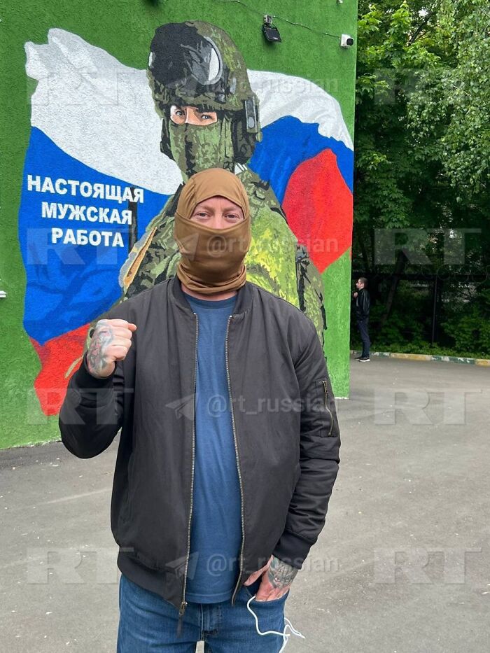 Man wearing a face covering and jacket standing with fist raised in front of a mural of a soldier and Russian flag.