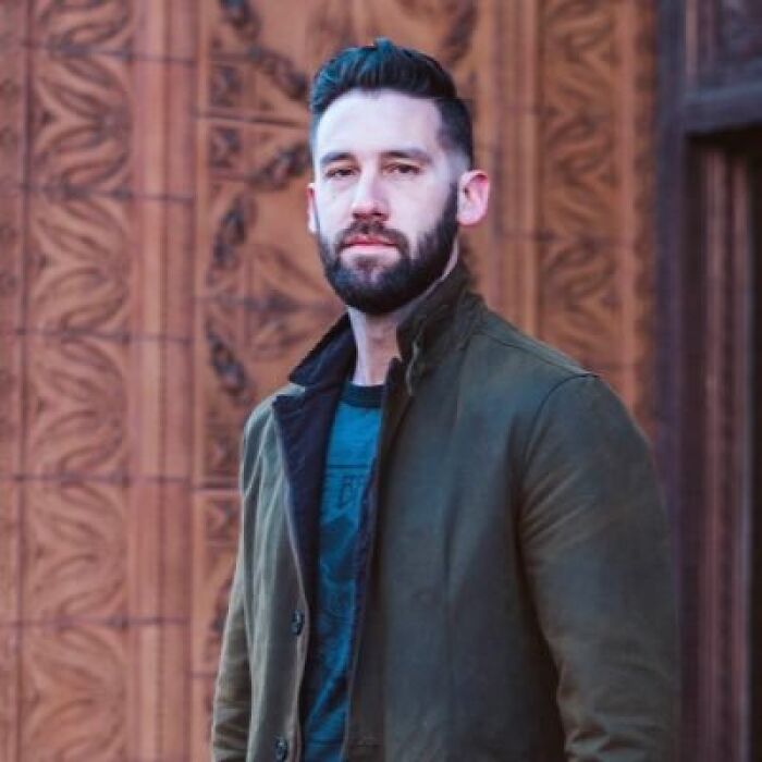 Man with beard and jacket standing outdoors in front of decorative wooden panel, related to Pulitzer winner cartoon controversy.