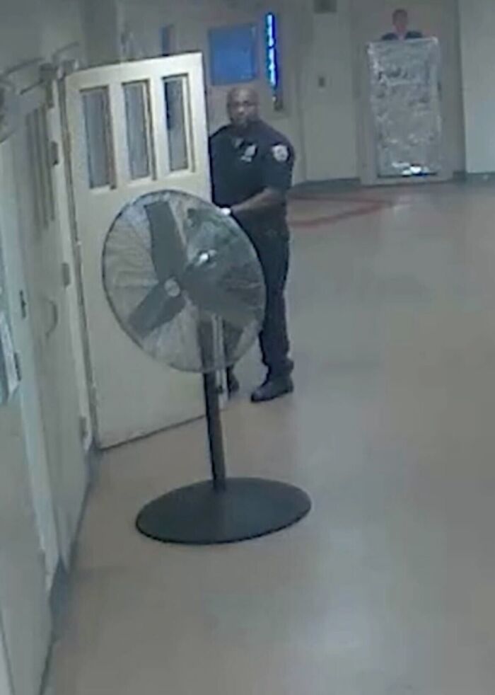 Security officer standing in a Rikers Island hallway near a large fan, highlighting systemic dysfunction in the facility.