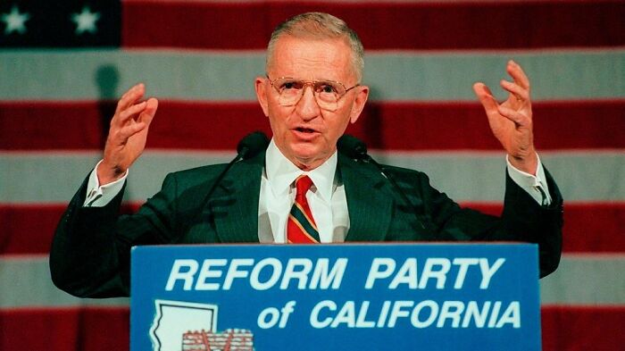A man in glasses and suit speaking at a Reform Party of California podium with an American flag backdrop.