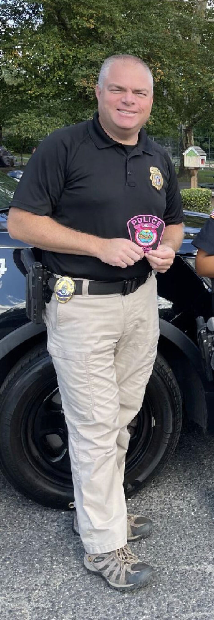NJ police chief in uniform holding a police patch outdoors near a police vehicle with trees in the background