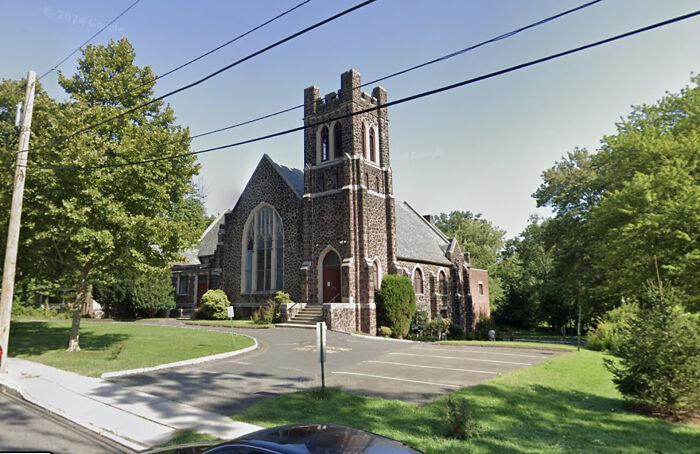 Stone church building with tower and arched windows under clear sky near trees and empty parking lot in NJ police chief abuse case area.