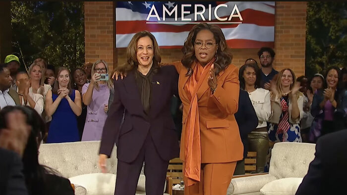 Kamala Harris and Oprah Winfrey on stage during a public event with an audience in the background and America flag banner.