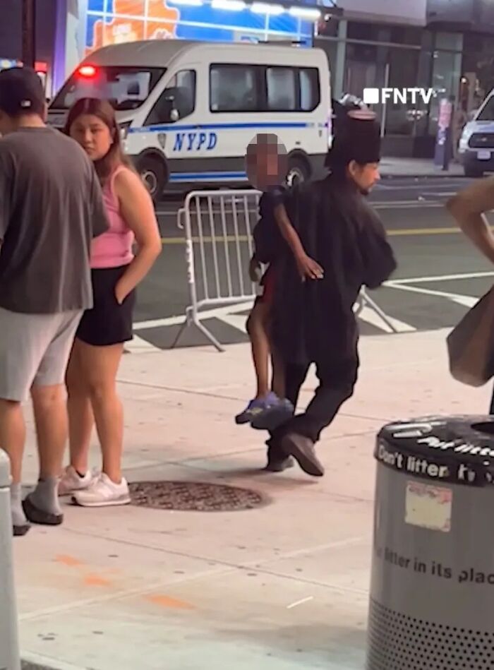 Man in black clothing lifting boy aged six at Coney Island with NYPD van and bystanders nearby on the sidewalk.