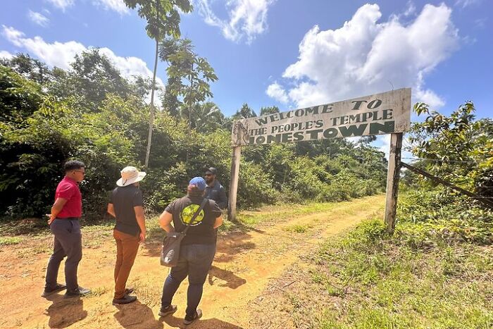 Visitors stand near a weathered Jonestown entrance sign surrounded by lush greenery under a partly cloudy sky.