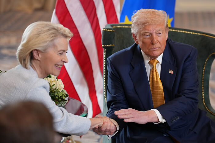 Donald Trump in a dark suit shaking hands with a woman, with American and European Union flags in the background.