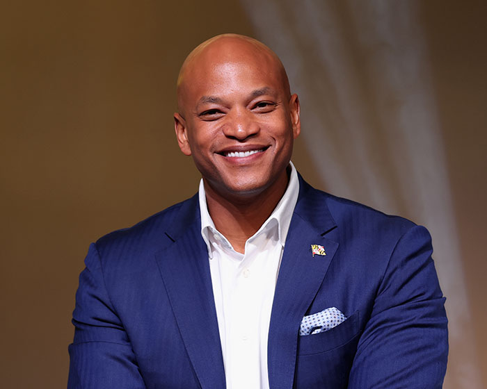 Smiling man in blue suit jacket and white shirt, posing confidently against a plain brown background.