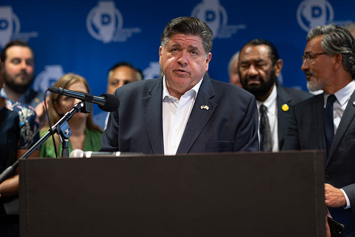 Democratic governor speaking at a podium with a microphone, surrounded by a diverse group of people behind him.