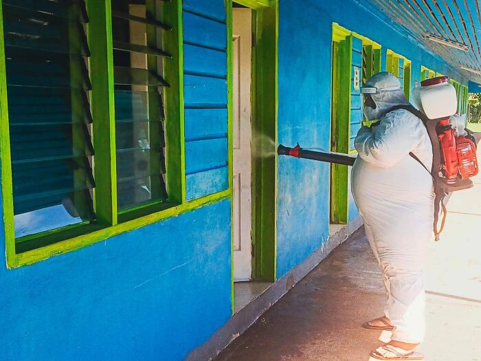 Person in protective gear spraying insecticide outside a blue building amid Pacific Islands dengue fever outbreak concerns.
