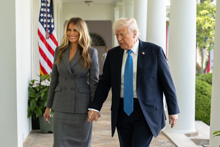 Melania Trump and Donald Trump walking hand in hand near American flag in a formal outdoor setting.