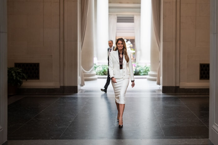 Melania Trump walking confidently in a white skirt suit in a grand hallway addressing Hunter Biden Epstein claims.