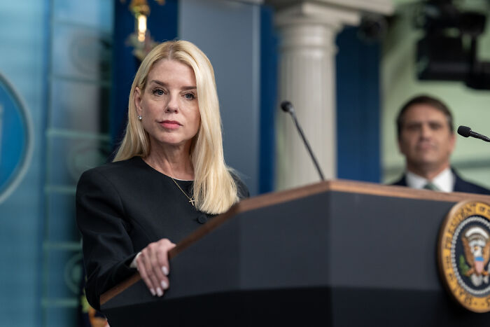 Woman standing at a podium with presidential seal, related to investigation of Sen. Adam Schiff mortgage fraud violations.