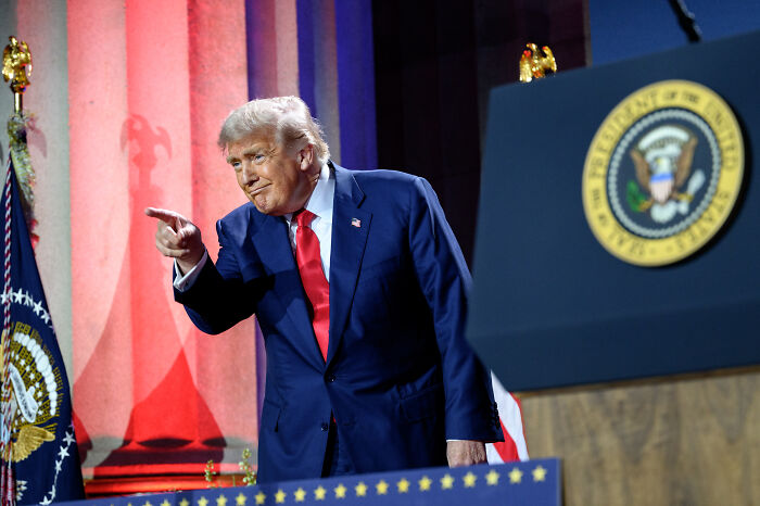 Donald Trump in a dark suit and red tie pointing during a speech, with United States presidential seal visible behind him.