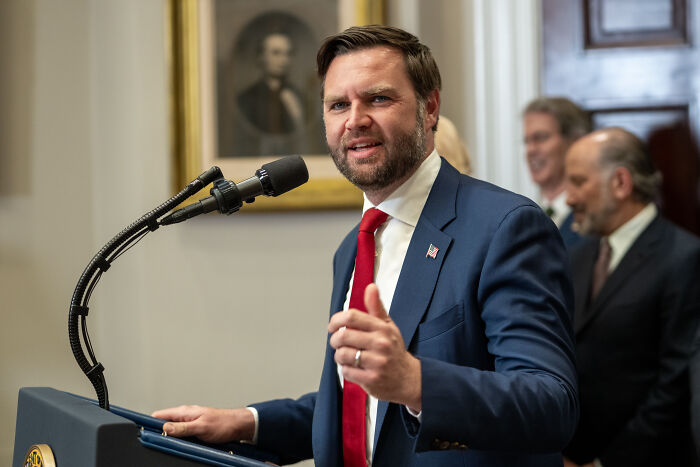 JD Vance speaking at a podium in a suit and red tie, addressing an audience during a formal event indoors.