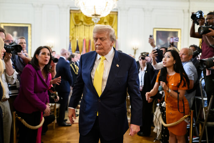 Donald Trump walking in a crowded room with reporters and cameras, discussing National Guard troops deployment plans.