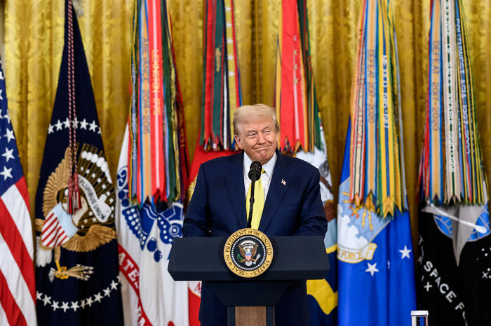 Former President Trump in a suit speaking at a podium with flags behind, linked to Alaska and Russia confusion before Putin meeting