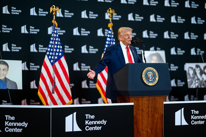Donald Trump speaking at The Kennedy Center podium with American flags in the background on leadership and dictatorship claims.