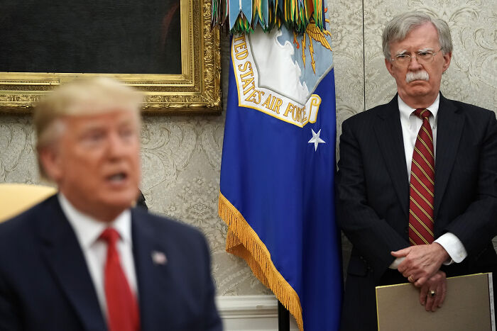 John Bolton standing in a suit near United States Air Force flag during major national security FBI investigation.
