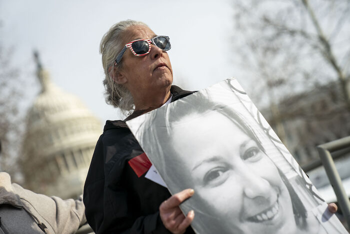 Woman wearing patriotic sunglasses holding a photo near Capitol, related to Jan 6 Capitol rioter receiving military funeral honors.