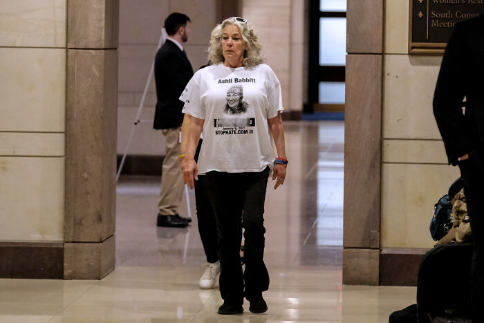 Woman wearing a shirt with Ashli Babbitt image walking inside a government building related to Jan 6 Capitol rioter news.