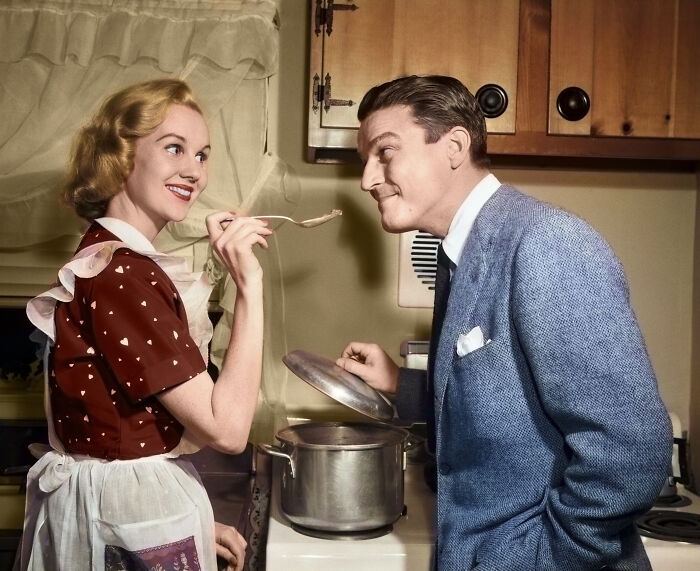 Woman in vintage dress and apron feeding man in suit in a kitchen, illustrating tradwife concept in modern political climate.