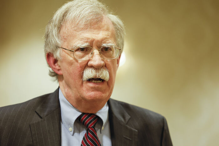 John Bolton speaking at an event, wearing glasses, a suit, and a striped tie amid national security investigation.