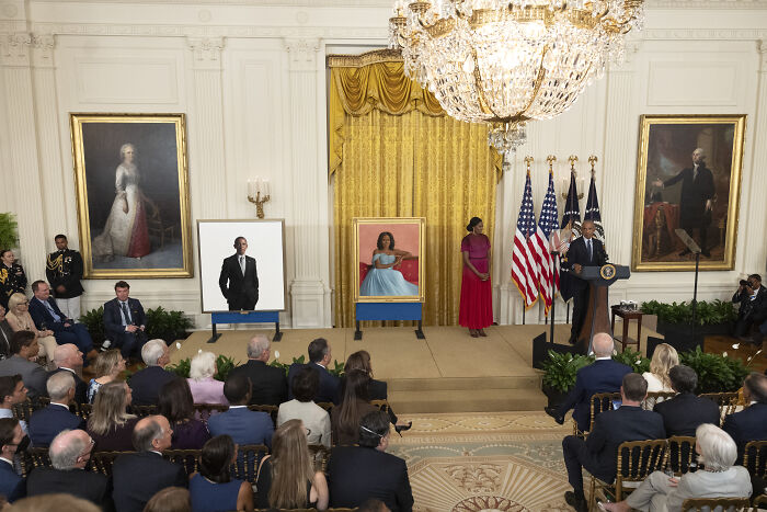 Barack Obama portrait displayed in White House with audience and speaker, highlighting Trump moving the portrait to a concealed area.