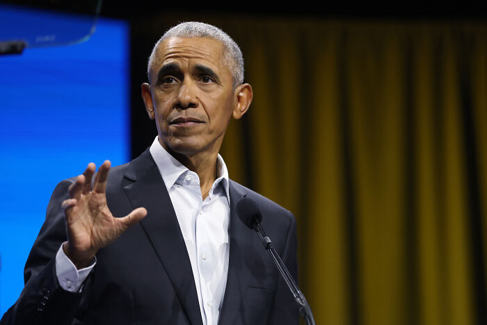 Former President Barack Obama speaking at a podium with a yellow curtain background, related to intelligence subversion clearance.