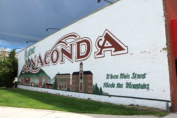 Mural of Anaconda historic district painted on a white brick wall with grass and cloudy sky, related to Montana bar shooting manhunt.