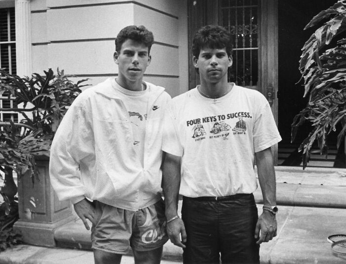 Erik Menendez and his brother standing outside a building, wearing casual 1990s clothing in a black and white photo