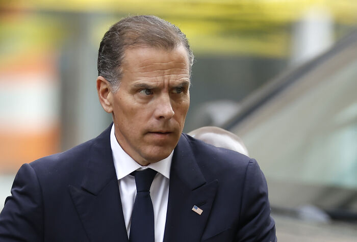 Hunter Biden in a dark suit with an American flag pin, looking serious while walking outdoors in an urban setting.