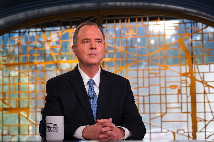 Adam Schiff seated at a desk with a coffee mug, wearing a suit and tie, in a news interview setting.
