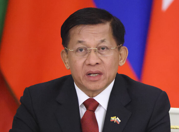 Senior Myanmar official speaking at a formal event, with flags in the background, amid sham election and atrocity allegations.