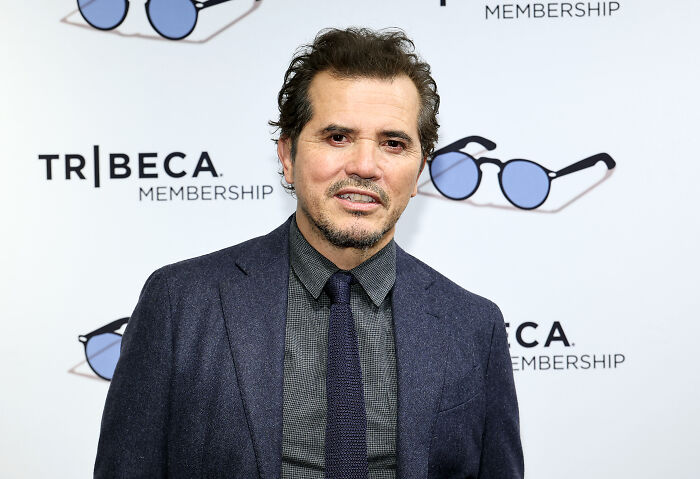 John Leguizamo at an event, wearing a dark suit and tie, with a Tribeca Membership backdrop behind him.