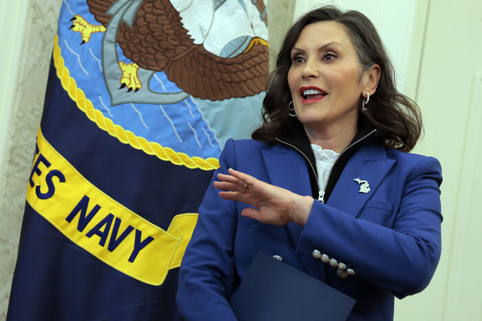 Woman in blue blazer speaking in front of a United States Navy flag, related to Trump and imaginary governor topic.