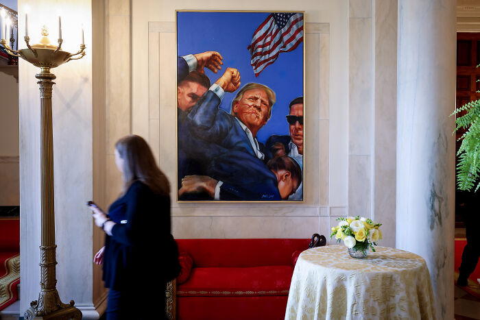 Portrait of Donald Trump with American flag displayed in White House with woman walking nearby, referencing Obama's concealed portrait move.