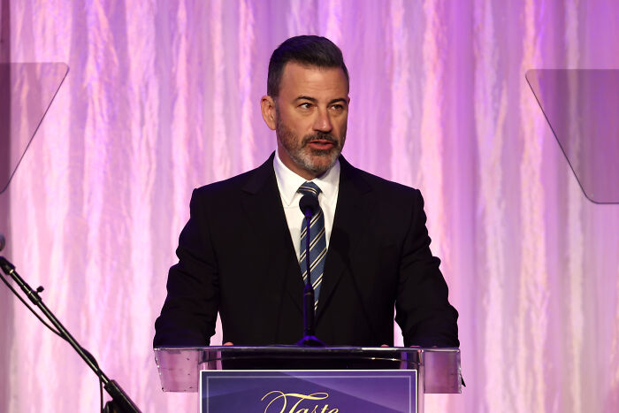 Jimmy Kimmel at a podium speaking, wearing a suit and tie, relating to new citizenship and celebrities leaving the U.S.