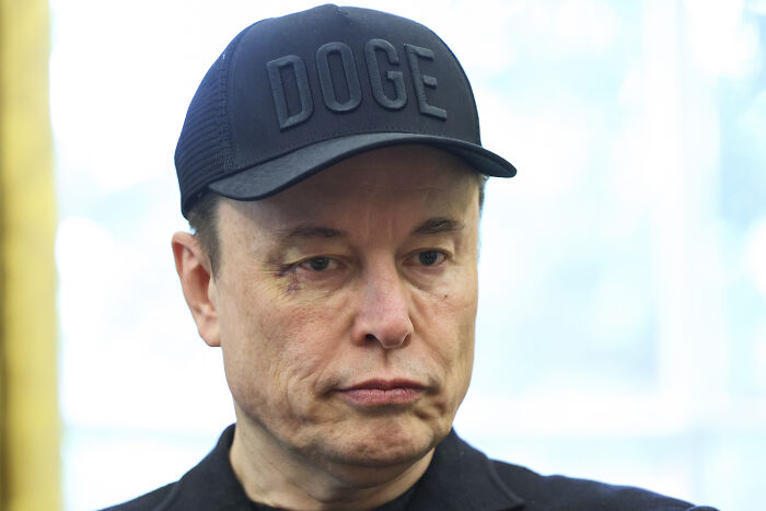 Man wearing a black DOGE cap, appearing serious while experts warn about complexity of large language models and AI orders.