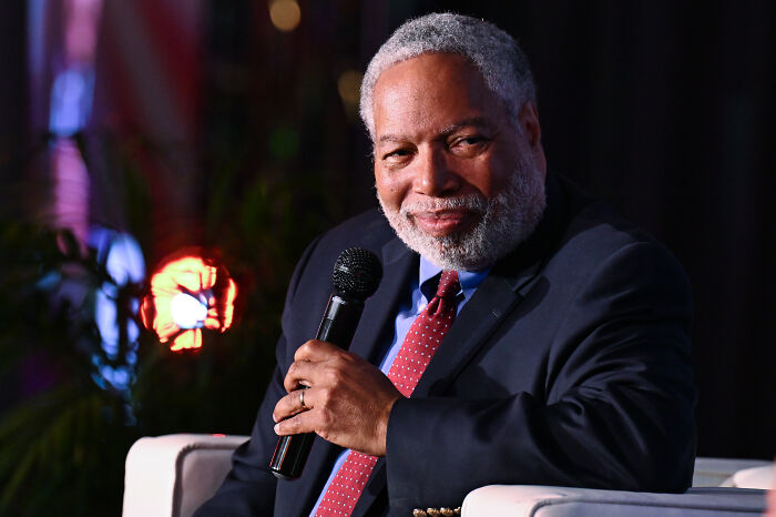 Man with gray hair and beard in suit holding microphone, speaking at event about Trump slams Smithsonian on slavery.