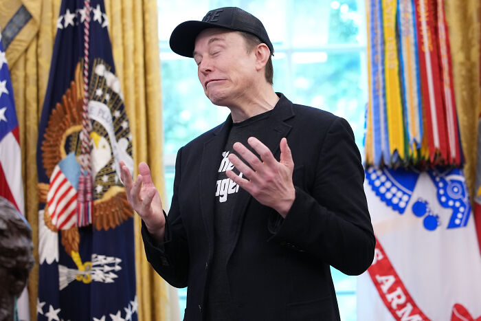 Man gesturing with hands in a political setting featuring flags and symbols related to Skibidi, Delulu, Tradwife, and Broligarchy.