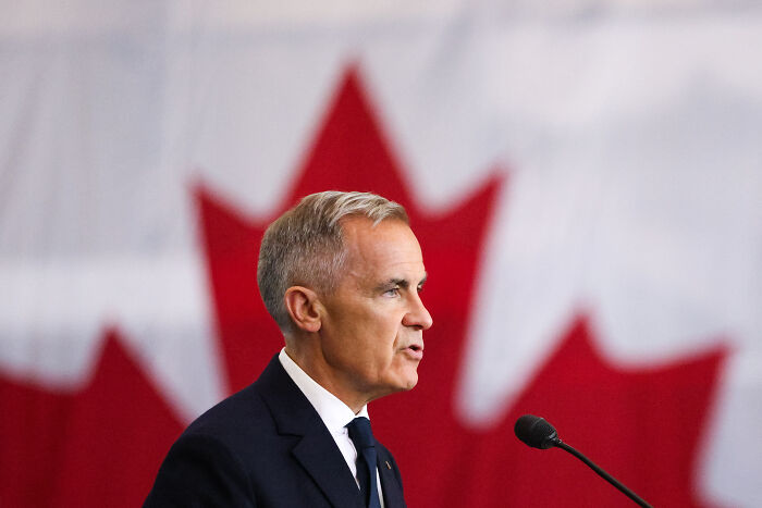 Canadian official speaking at a podium with a blurred Canadian flag in the background amid tariff tensions with the US.