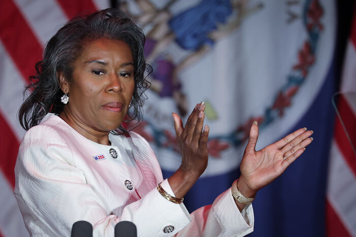 Woman in white suit clapping during event with US and Virginia state flags in background about trans rights protester outrage