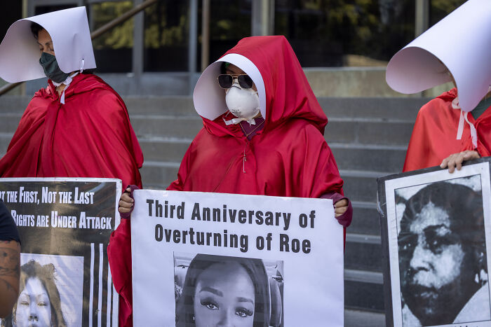 Protesters dressed as handmaids holding signs on the third anniversary of overturning Roe highlighting political climate keywords.